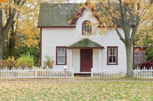 Fenced in white house with red door, surrounded by a few trees in the fall